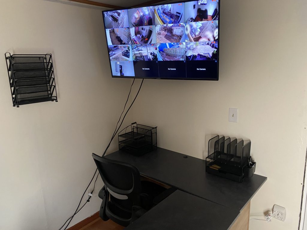 Office corner with a wall-mounted monitor displaying multiple CCTV feeds, a desk with an office chair, and document organizers.