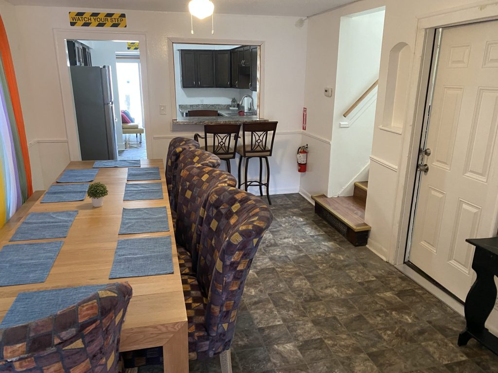 Dining area with patterned chairs and a wooden table, view of kitchen through a window, and stairs near the door in a cozy interior.