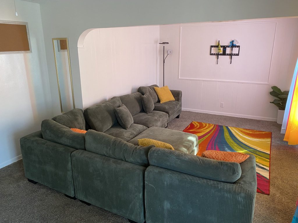 A green L-shaped sofa with yellow and orange cushions, a colorful rug, and a small wall shelf in a sparsely decorated room.