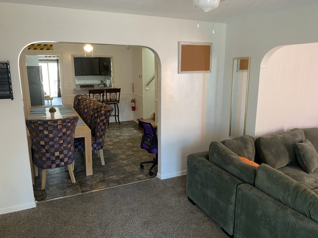 Open living space with a green sofa, dining table with patterned chairs, and a view into the kitchen with a purple swivel chair.